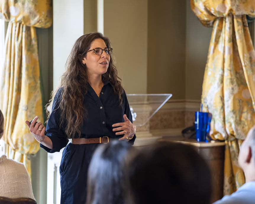 Woman with glasses speaking in front of an audience in a room with yellow curtains.