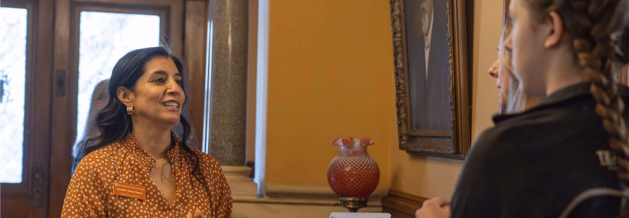 Woman in orange polka dot blouse talking to two other women in a room with framed art and a decorative vase.