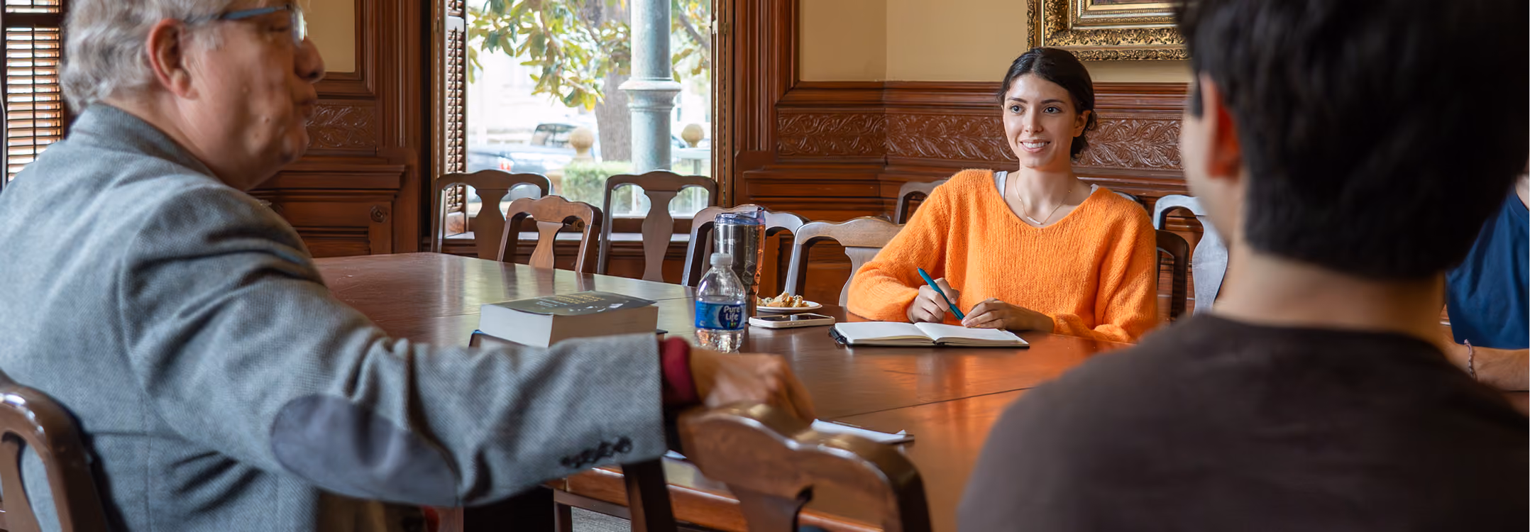 Young woman in orange sweater smiles while writing in a notebook at a large conference table.