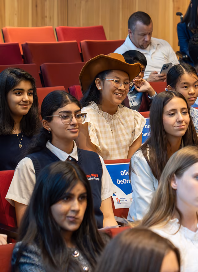 A diverse group of young women seated in red auditorium chairs, some smiling and attentive.