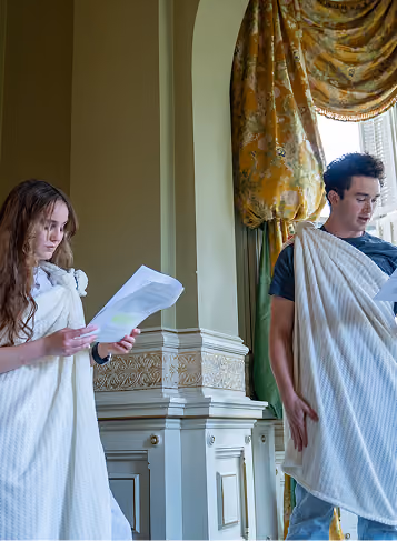 Two people in togas reading papers in a decorated room with ornate walls and curtains.