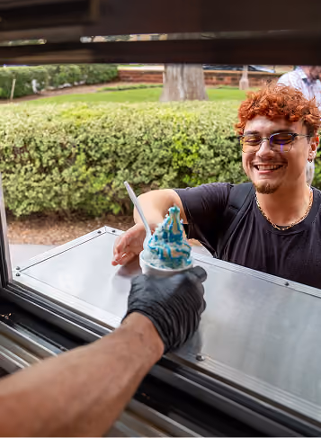 Smiling man with glasses receives a blue and white ice cream cone from a gloved hand at a service window.