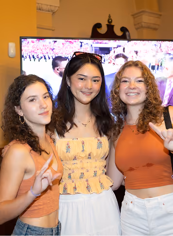 Three smiling young women posing indoors with two making peace signs and a large TV behind them.