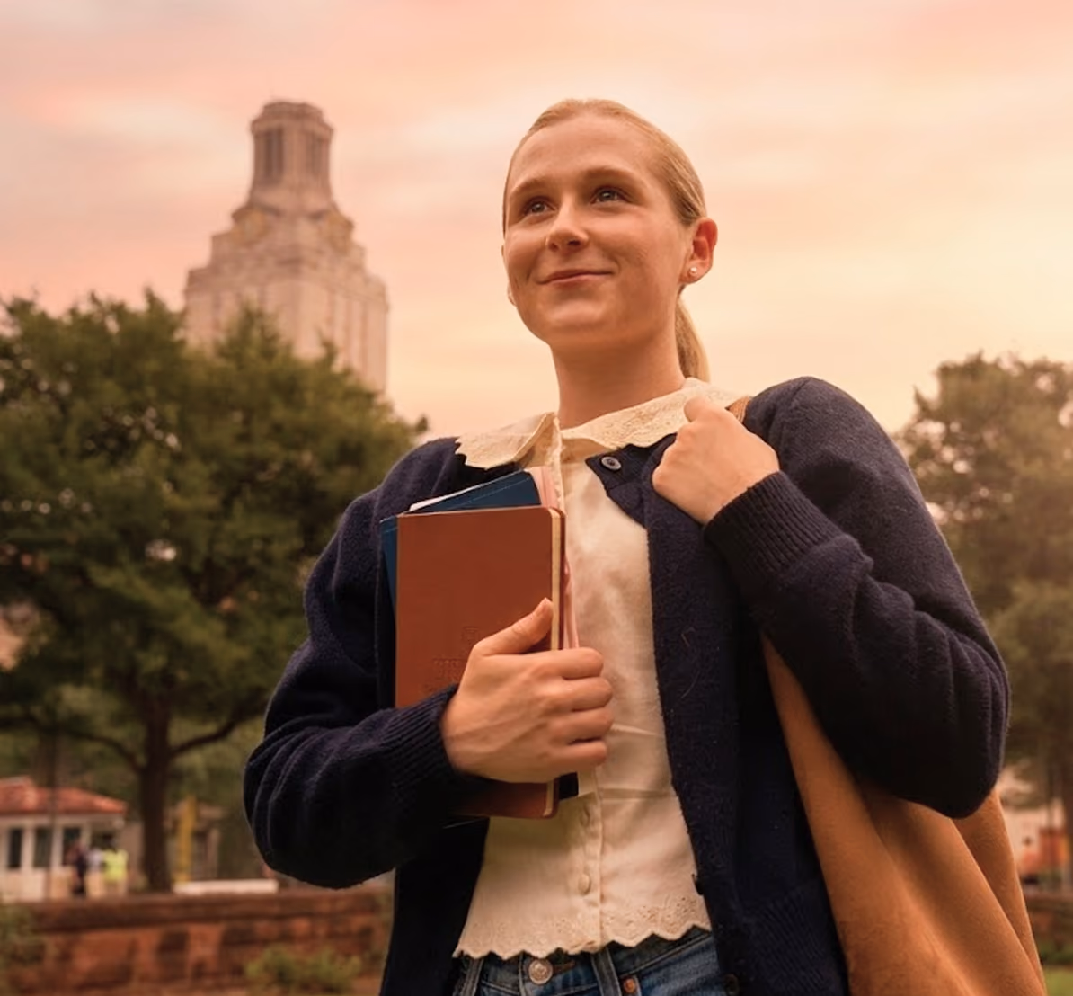 Young woman with books and tote bag outdoors in front of a tower at sunset.