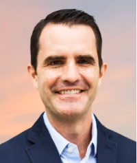 Smiling man with short dark hair wearing a dark blazer and light shirt against a soft sky background.