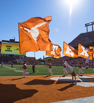 College football players run on field waving Texas Longhorns flags under bright sun.