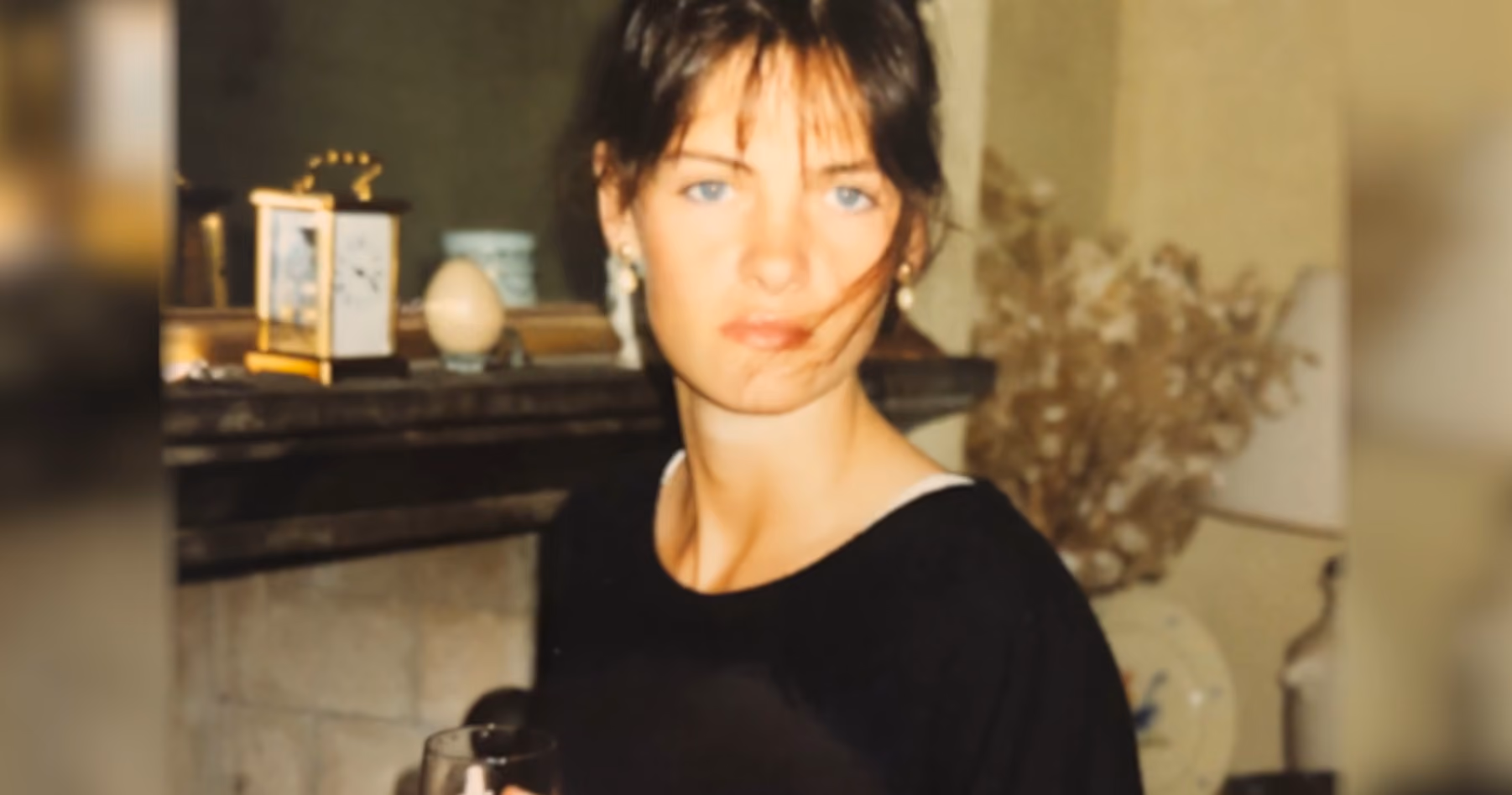 Young woman with blue eyes and dark hair wearing a black top holding a glass indoors.