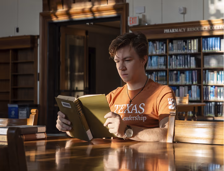 Young man in an orange Texas Leadership shirt reading a book in a sunlit library.