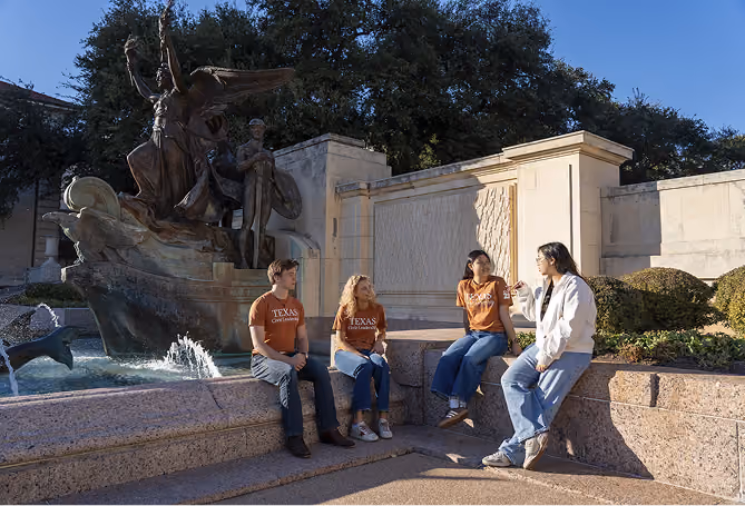 Four students sitting near a large fountain sculpture outside on a sunny day, talking and relaxing.