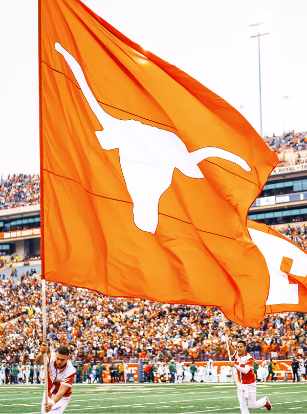 Two people running on a football field holding large orange flags with white longhorn logos.