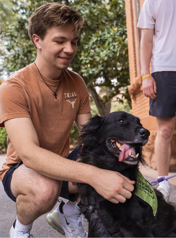 Young man in Texas shirt smiling and petting black service dog with green vest outside.