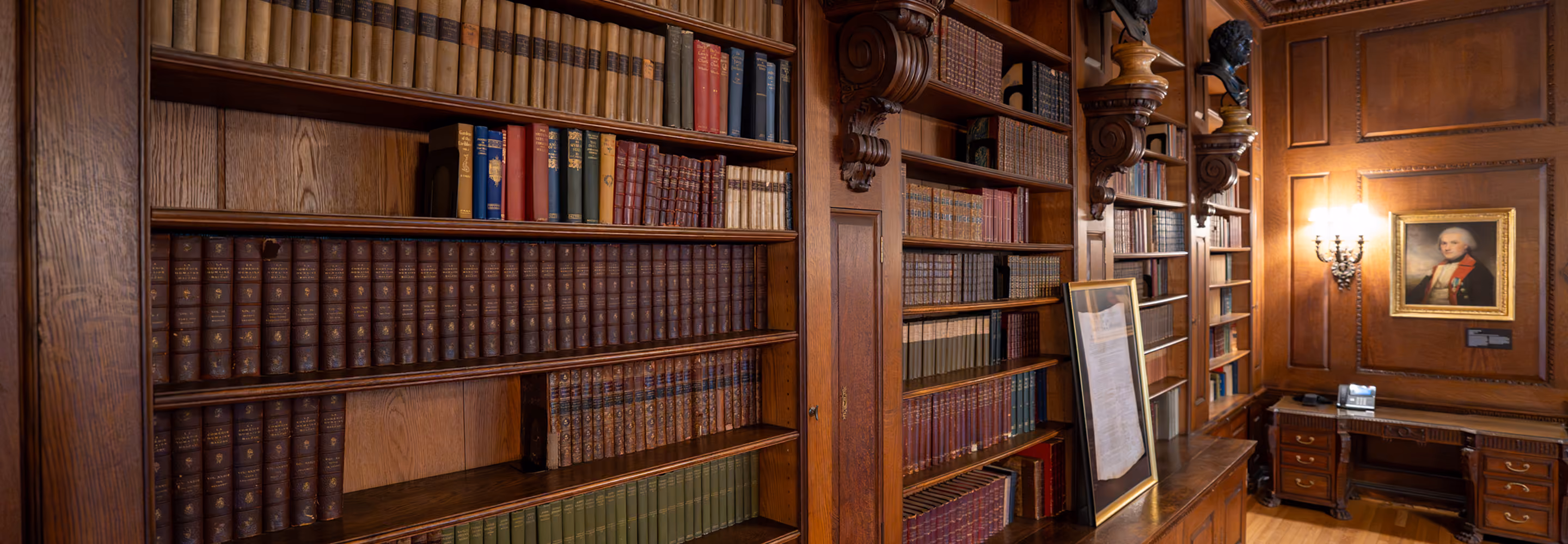 Wood-paneled library with shelves of old books, a framed document, desk, and a portrait on the wall.