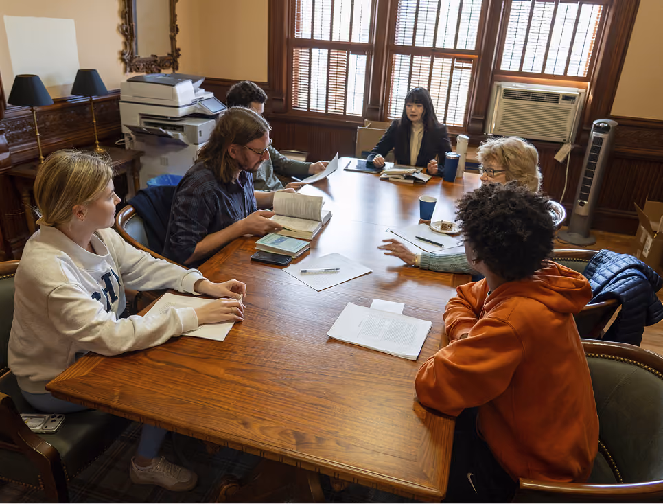 Six people sitting around a wooden table in a room, reading and discussing documents.