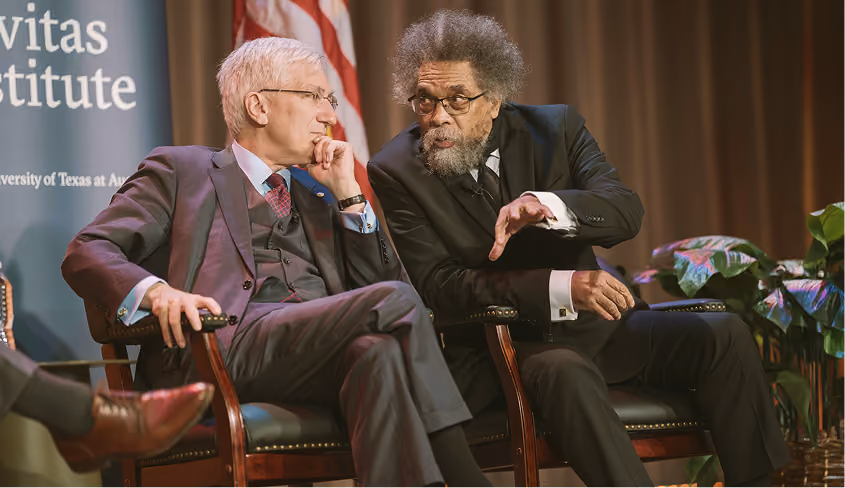 Two men in suits engaged in a serious discussion while seated on stage chairs.