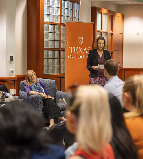 Woman standing and reading a paper near Texas Civic Leaders banner as audience listens in a room.