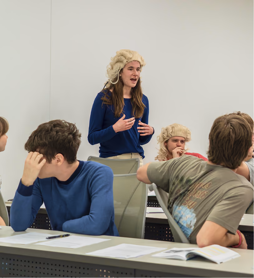 Students in a classroom, one standing and speaking wearing a white curly wig, others seated and listening.