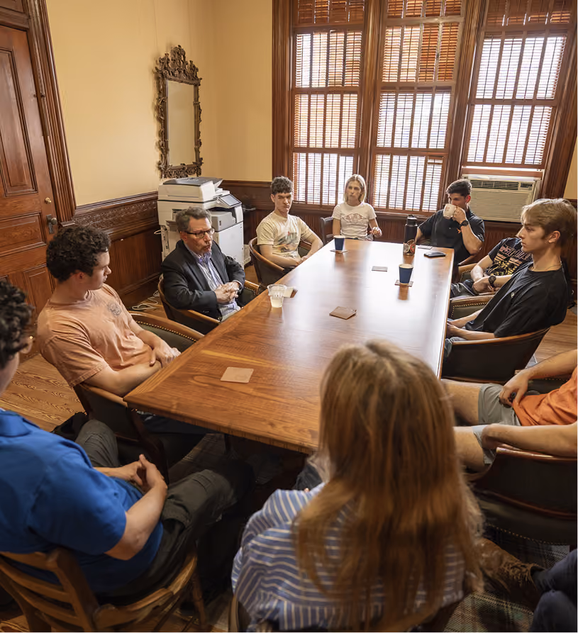 Group of people seated around a wooden table in a room with large windows and wood paneling.