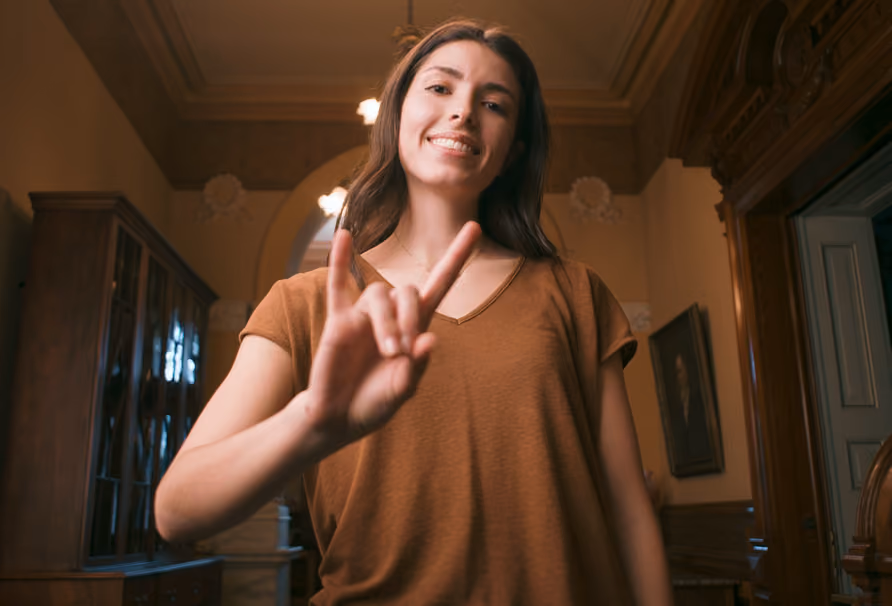 Smiling woman in brown shirt showing the 'I love you' sign with fingers indoors.