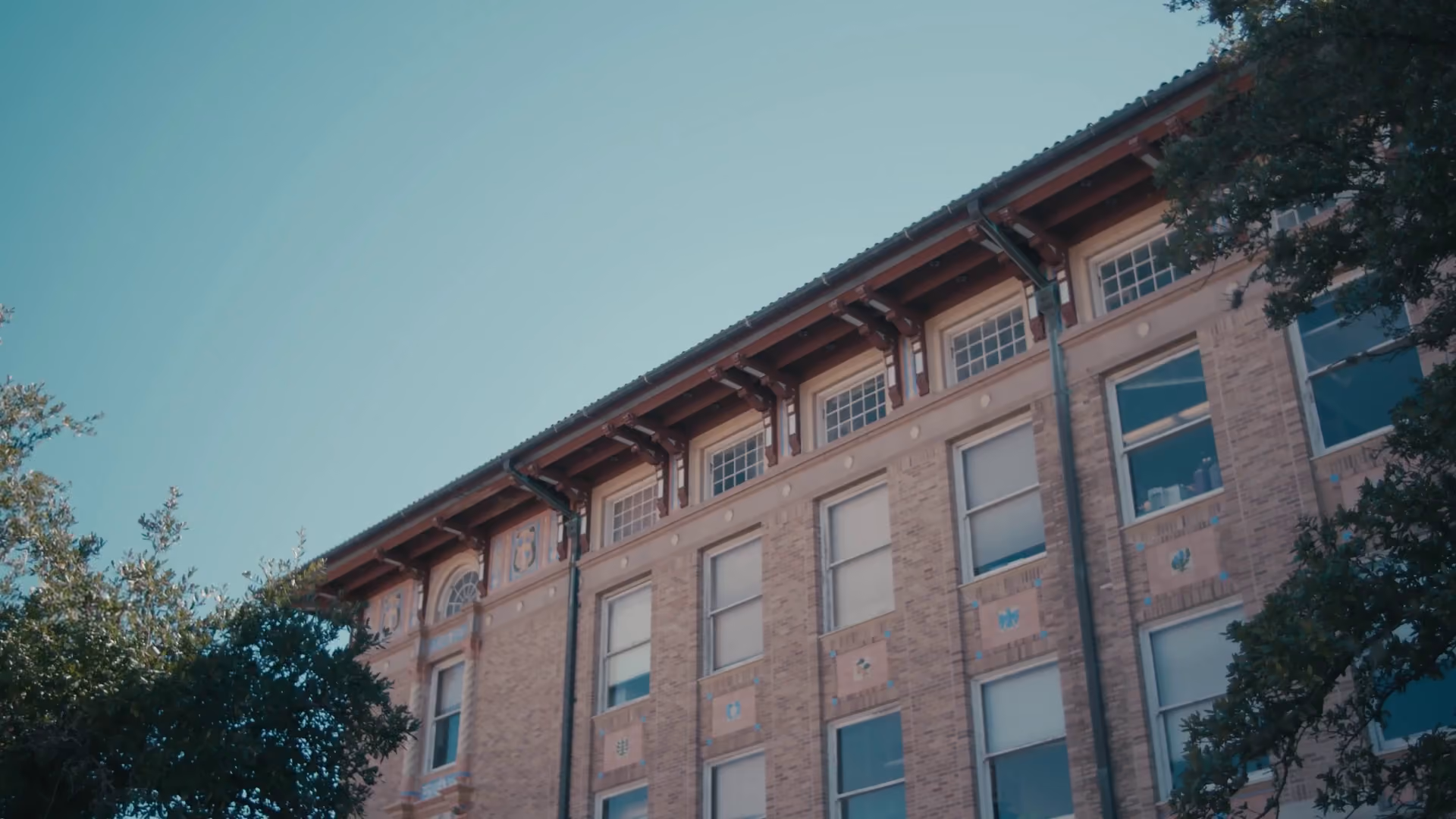 Facade of a tall brick building with multiple windows under a clear blue sky and trees.