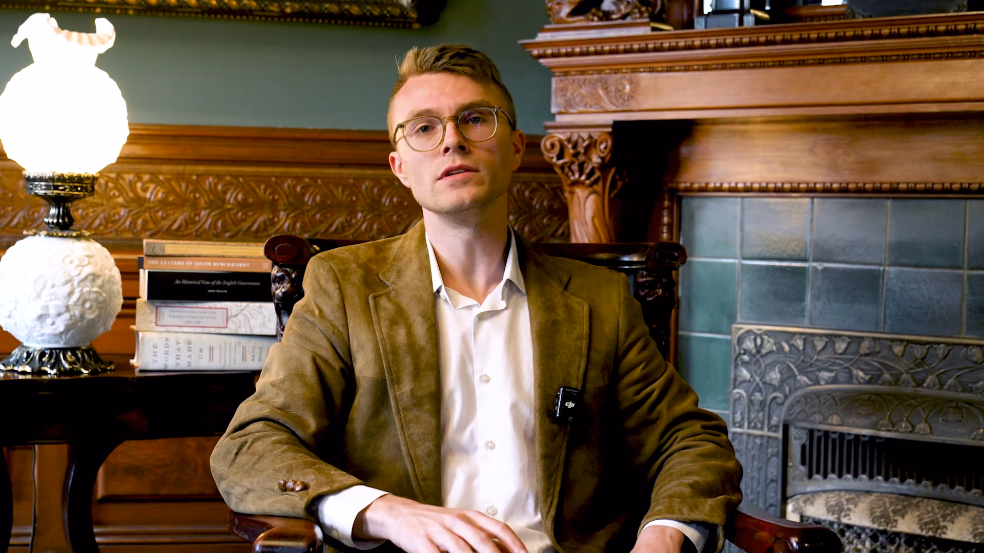 Man with glasses and brown jacket sitting in an ornate chair near a fireplace and a lamp.