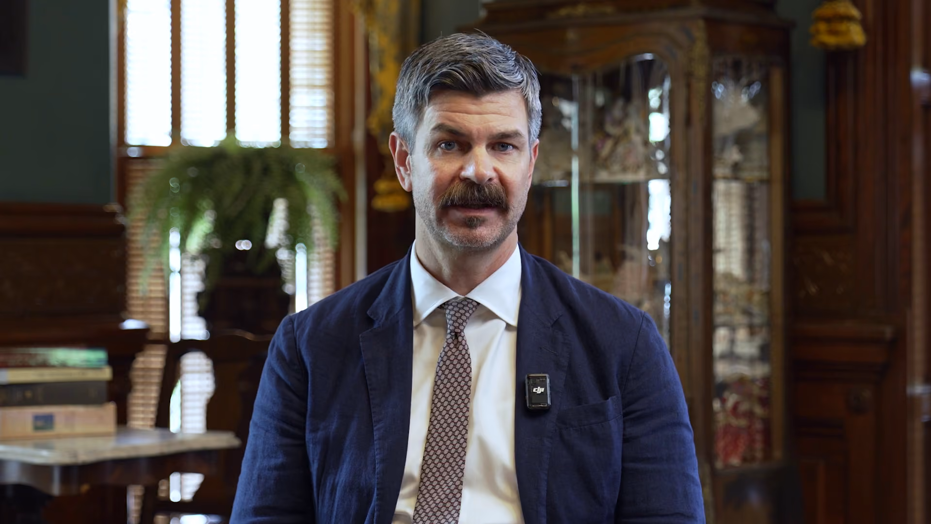 Man with gray hair and mustache wearing a blue jacket and patterned tie sitting indoors.