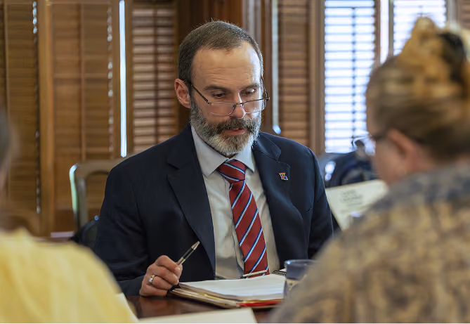 Man in a suit and striped tie reads documents at a table with others in a wood-paneled room.