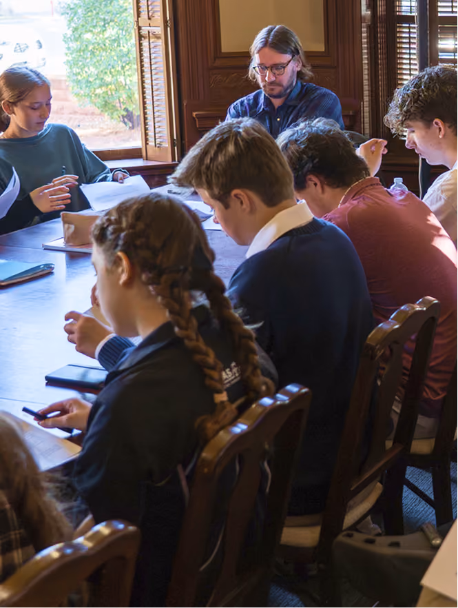 Students sitting around a table, focused on reading and writing in a room with wooden windows.