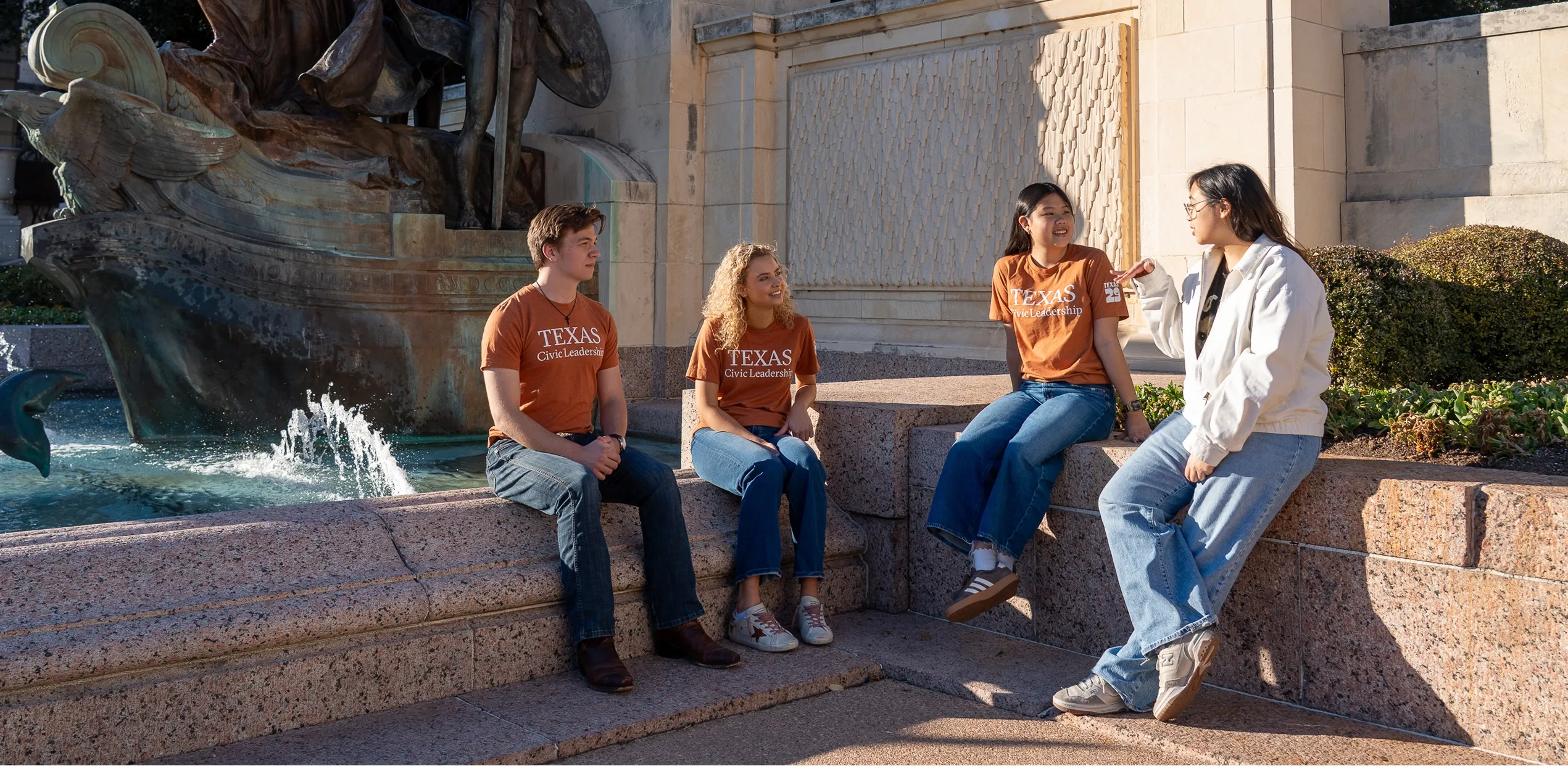 Four students sitting on stone steps near a fountain, three wearing Texas Civic Leadership shirts.