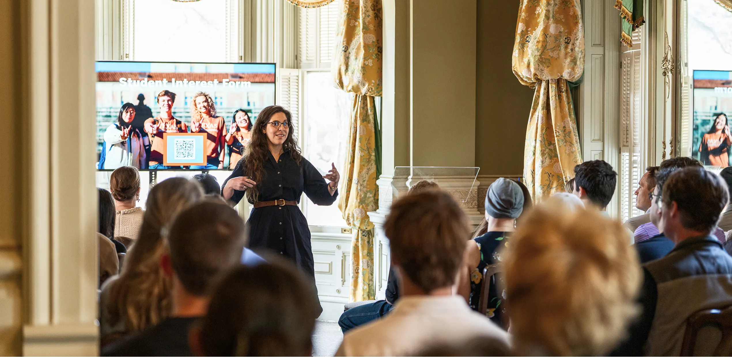 Woman giving a presentation to an audience in a room with large windows and yellow curtains.