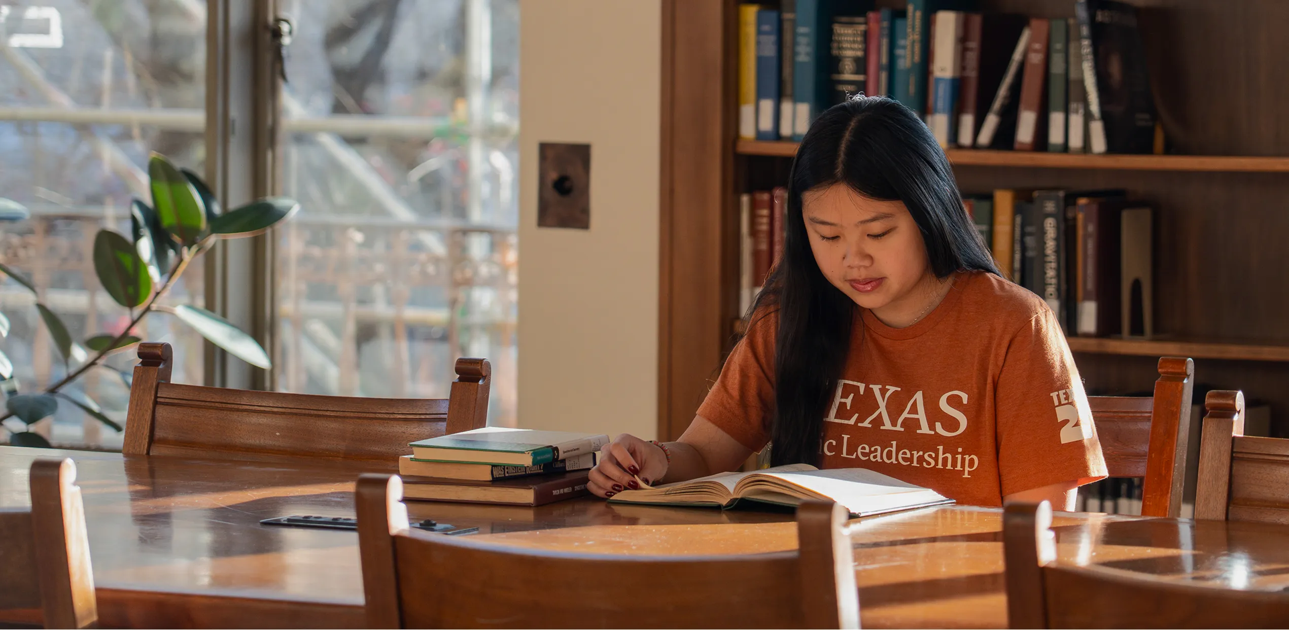 Young woman in Texas Leadership shirt reading a book at a library table with stacks of books.