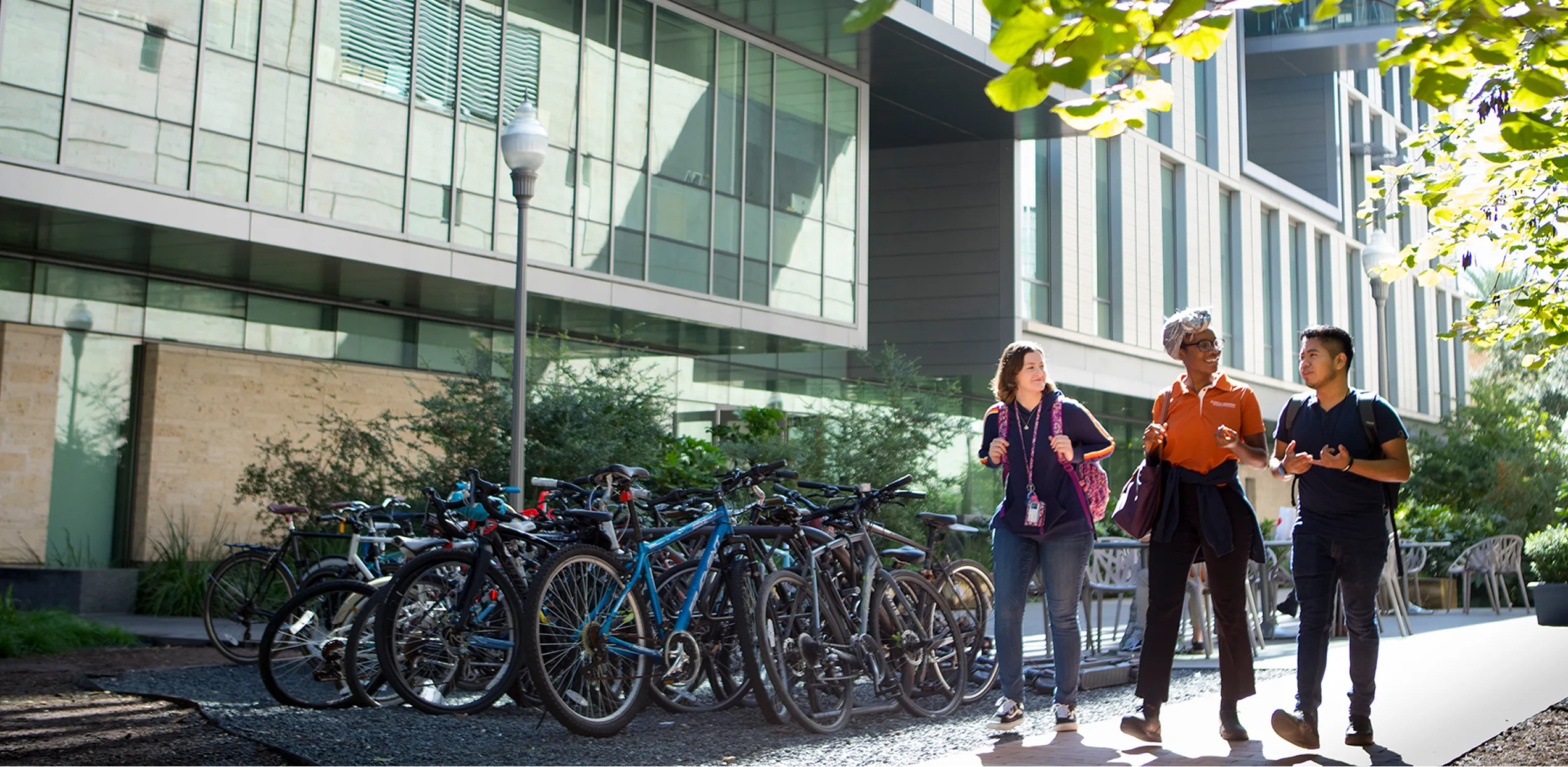 Three students with backpacks walking and talking near a bike rack outside a modern glass building.