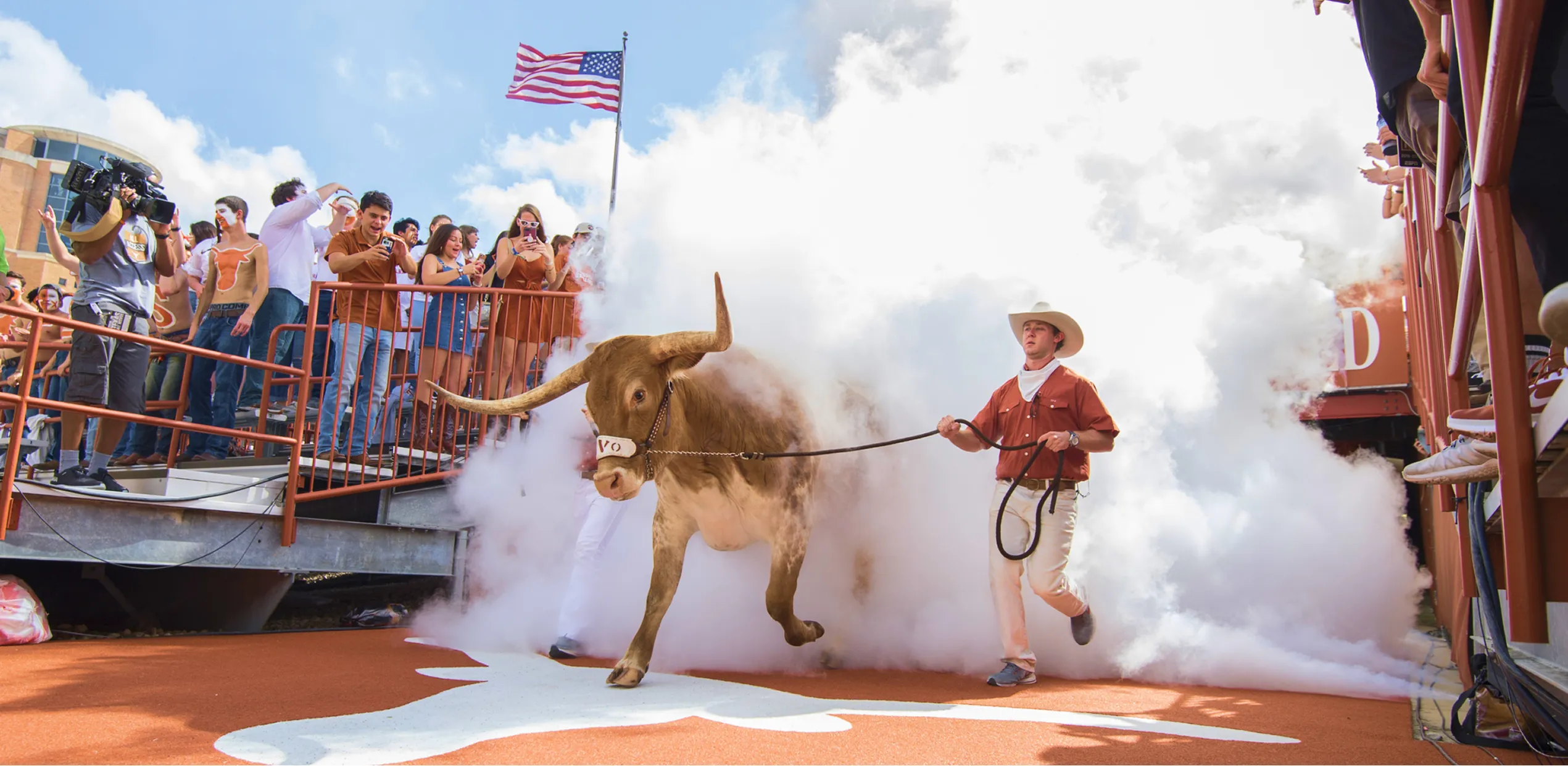 Longhorn steer led by a man in cowboy hat through smoke in front of cheering crowd and American flag.