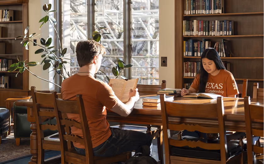 Two students reading books at a wooden table in a sunlit library with shelves and windows.
