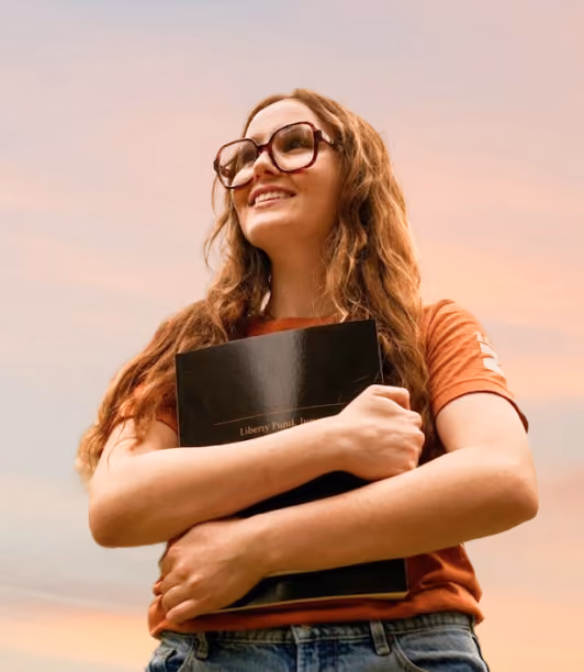 Smiling young woman with glasses holding a black book outdoors during a sunset.