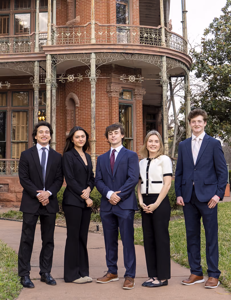 Five young adults dressed formally standing on a path in front of a large historic brick building with ornate iron railings.