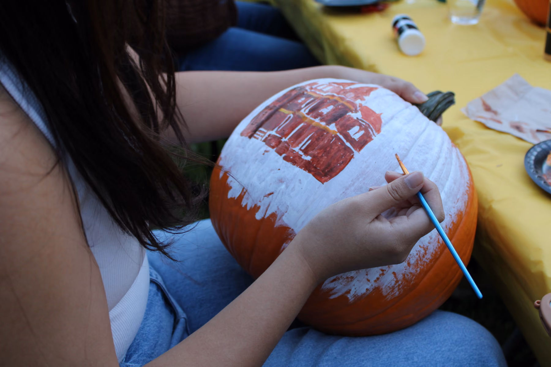Person painting a white base and red design of a building on a pumpkin with a brush.