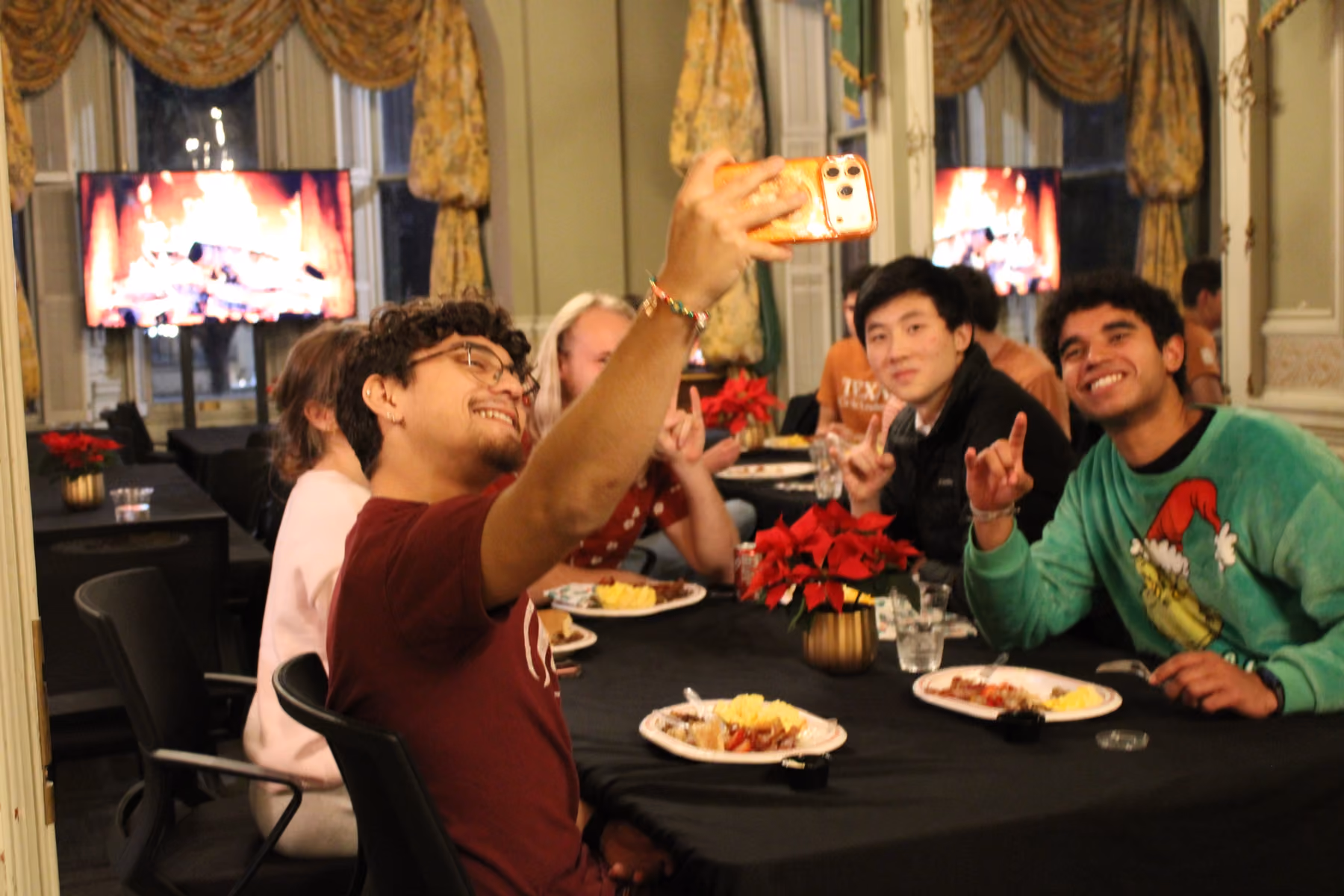 Group of young people at dinner table taking a selfie with a festive fireplace on TV behind them.