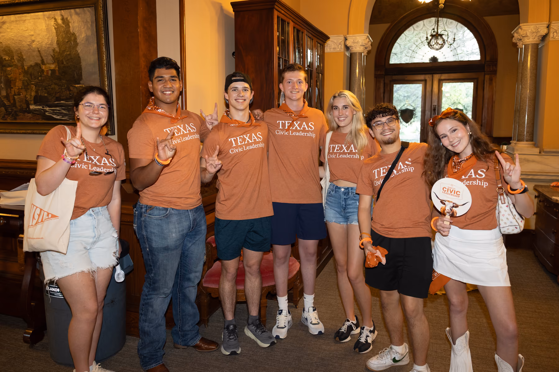 Seven young people in Texas Civic Leadership shirts posing indoors, smiling and making Hook ’em Horns hand signs.