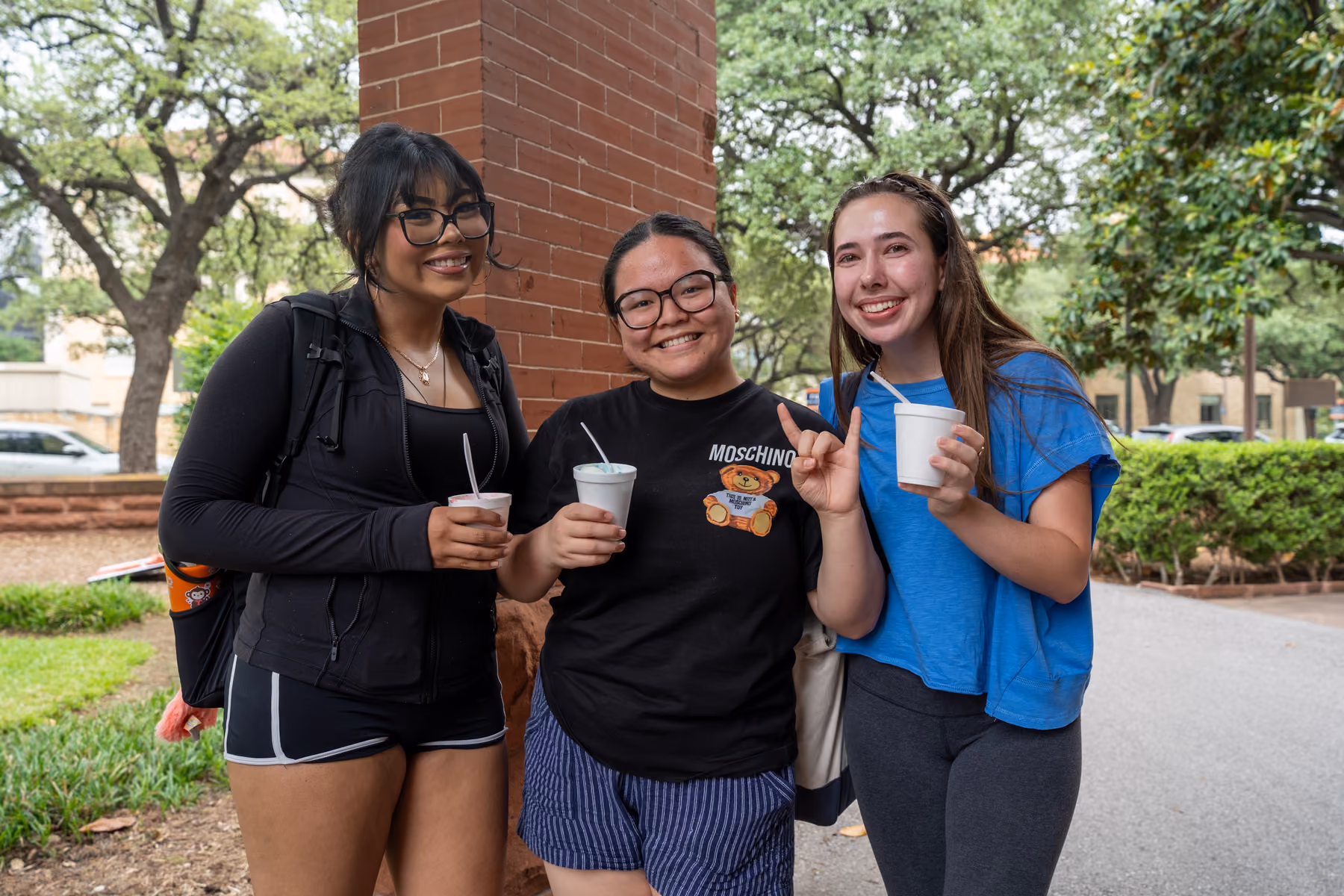 Three young women smiling outdoors holding cups with spoons near a brick pillar and trees.