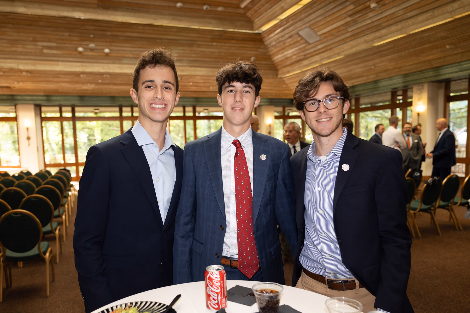 Three young men in suits smiling at a formal indoor event with chairs in the background.
