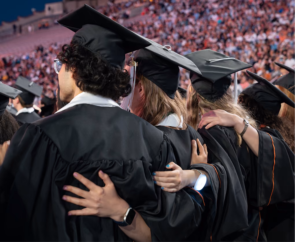 Group of graduates in black caps and gowns with arms around each other at ceremony.