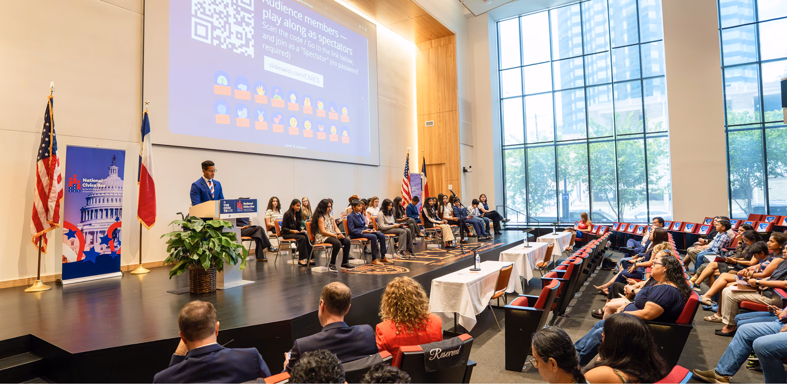 Youth at podium and seated on stage in a civic event room with audience watching in large glass-window hall.