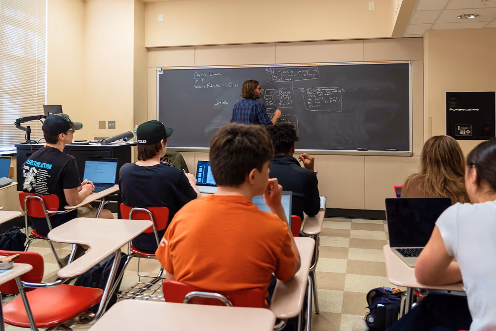 Students seated at desks in a classroom with laptops while a teacher writes on a chalkboard.