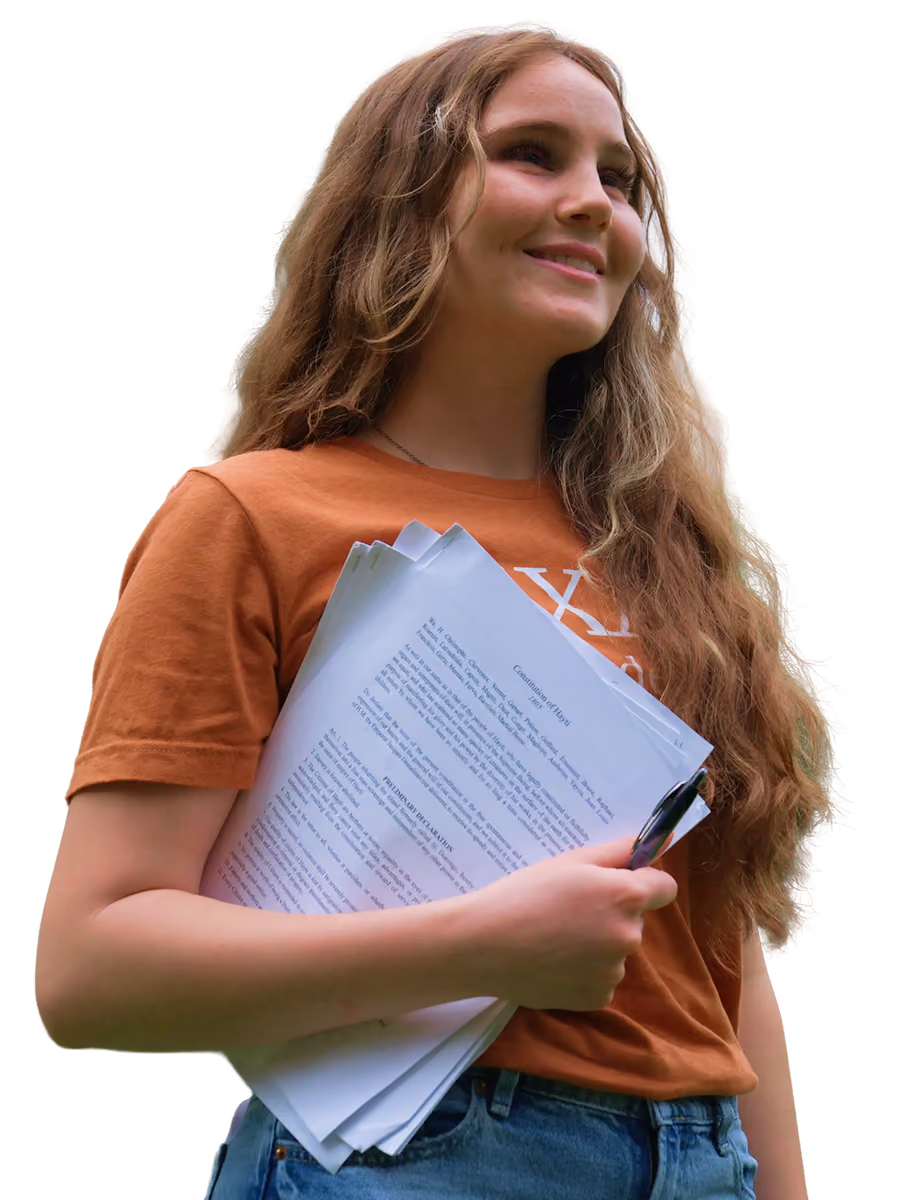 Smiling young woman with long hair holding papers and pen, wearing an orange shirt and jeans.