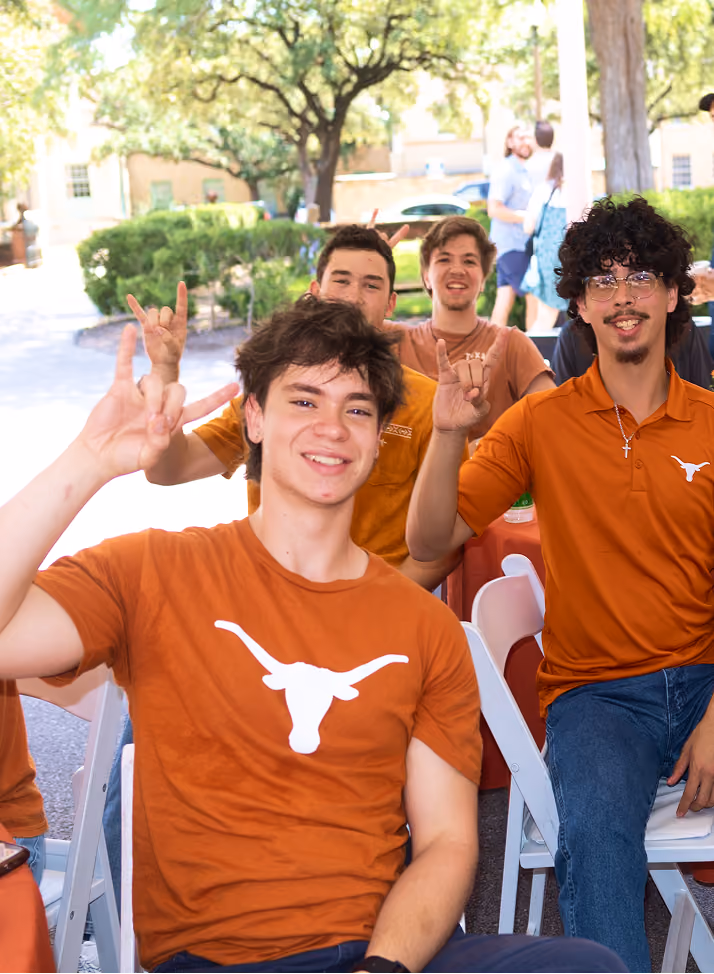 Young men wearing burnt orange Texas Longhorns shirts making hand signs while sitting outdoors.