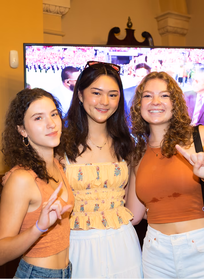 Three young women smiling indoors, two making hand gestures with index finger and pinky raised.