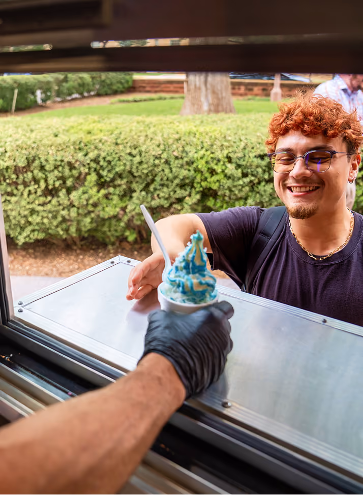 Smiling person with glasses receiving blue-topped soft serve ice cream through an open window.