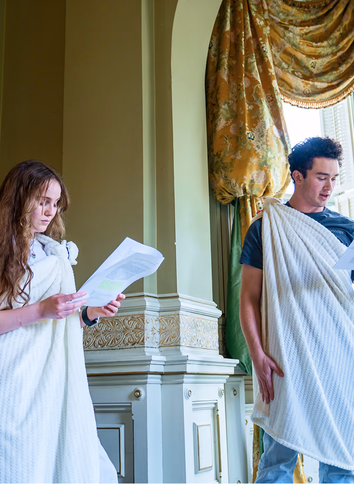 Two people wearing white togas reading from papers in an ornate room with green and gold curtains.