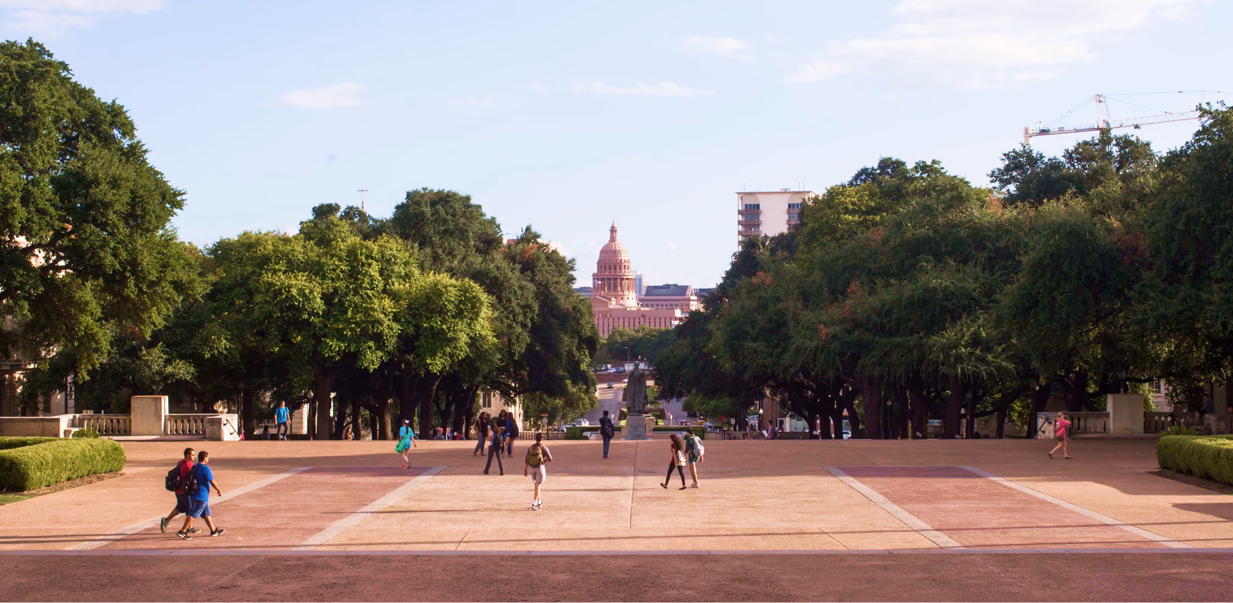 Wide walkway with trees on both sides and people walking toward a domed capitol building in the distance.