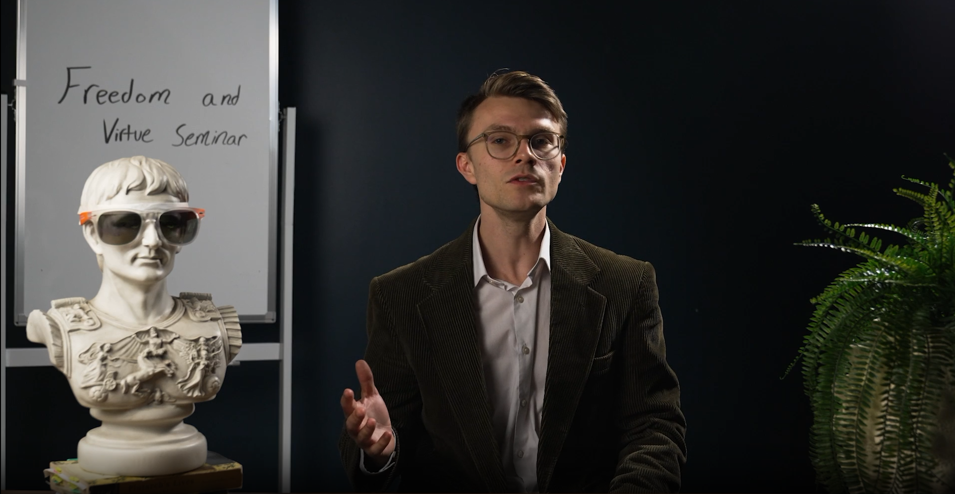 Man wearing glasses speaking in front of a bust with sunglasses and a whiteboard titled Freedom and Virtue Seminar.